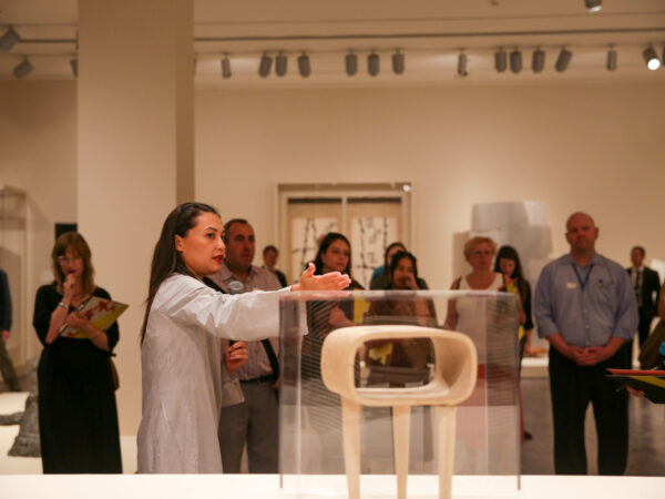 A curator gestures toward a contemporary sculpture in front of a crowd of visitors