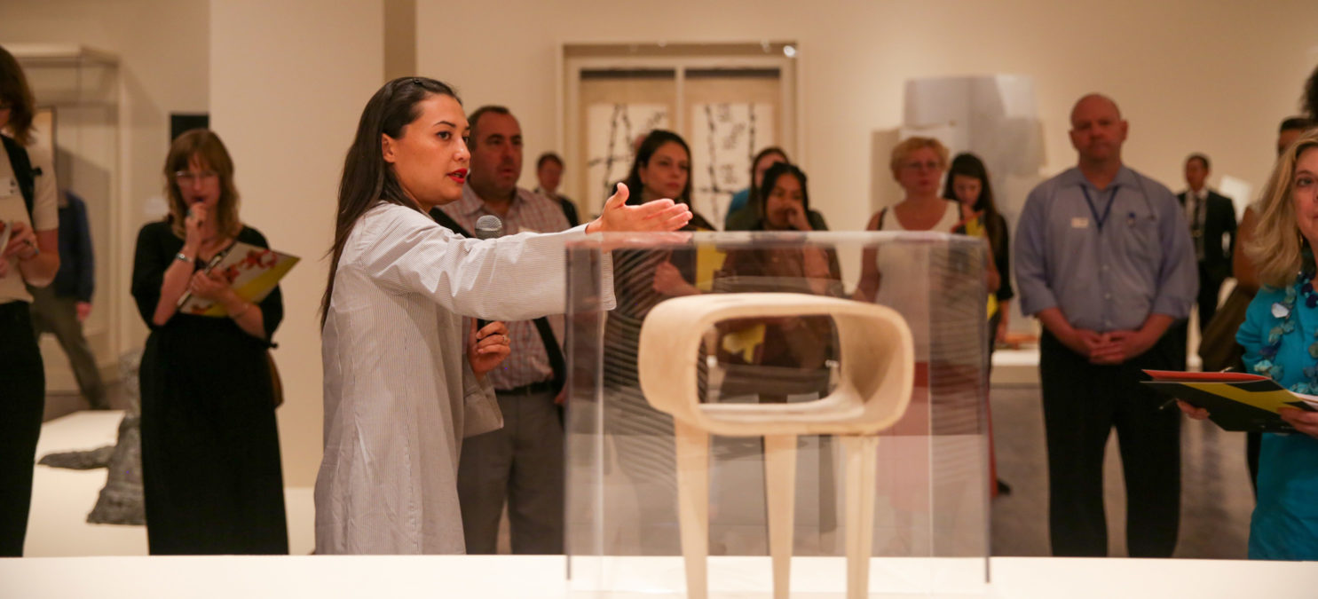 A curator gestures toward a contemporary sculpture in front of a crowd of visitors
