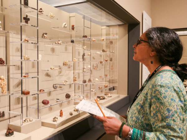 A woman looks at a large collection of netsuke in a glass case while holding large print descriptive labels.