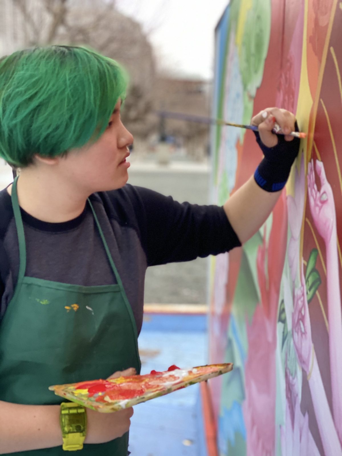 An artist with short green hair paints a colorful mural.